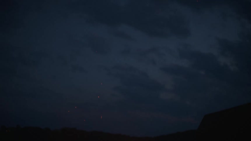 Sparks and embers from a bonfire drift upwards into the dark evening sky