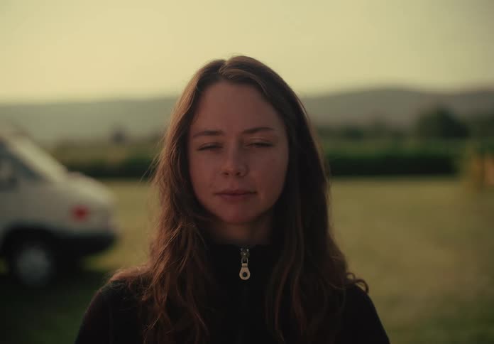 A serene young woman stands outdoors at sunset, opens her eyes, and stares into the camera