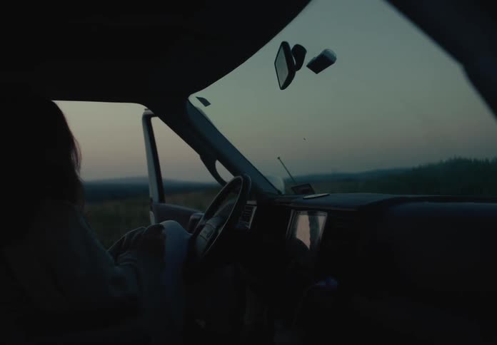 A solitary woman sits quietly in the driver's seat of a parked van in a rural field as twilight descends