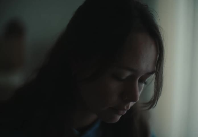 A somber close-up of a young woman's face in a dimly lit room, conveying sadness and deep contemplation