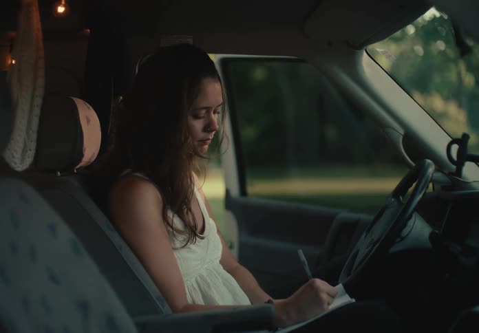 A thoughtful young woman sits in the driver's seat of her van, writing or drawing in her journal during a quiet moment on her travels