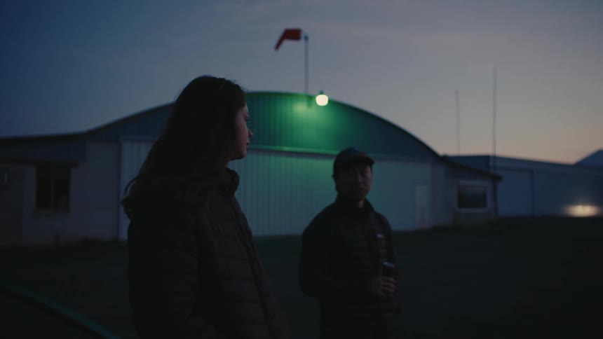 A man and a woman stand together quietly outside an airplane hangar at dusk, sharing a contemplative moment