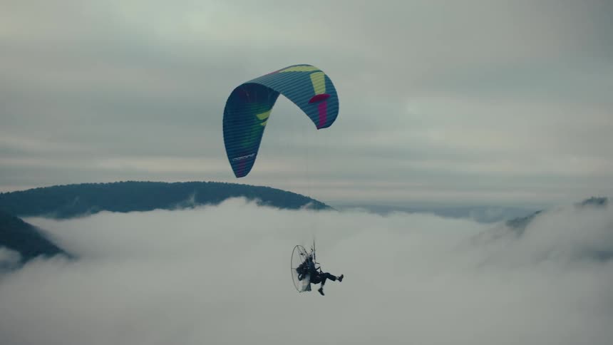 A person on a motorized paraglider soars peacefully above a thick blanket of clouds covering a mountain valley