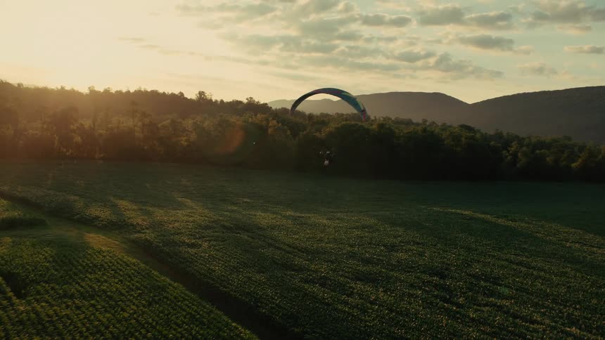 A powered paraglider flies low over a vast green agricultural field during a beautiful golden sunset