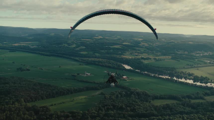 A breathtaking aerial shot follows a powered paraglider as it glides serenely over a vast, verdant valley and river