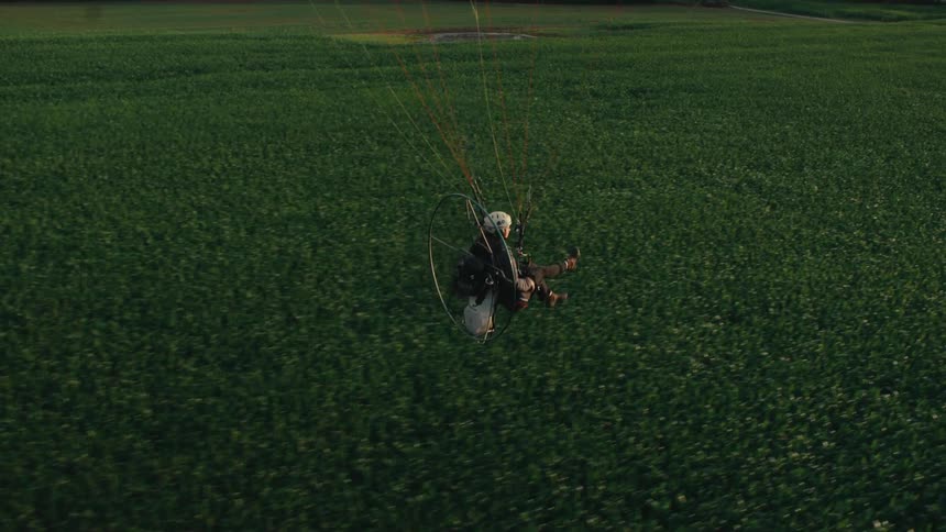 A serene aerial shot tracks alongside a paramotor pilot soaring over lush green farmland