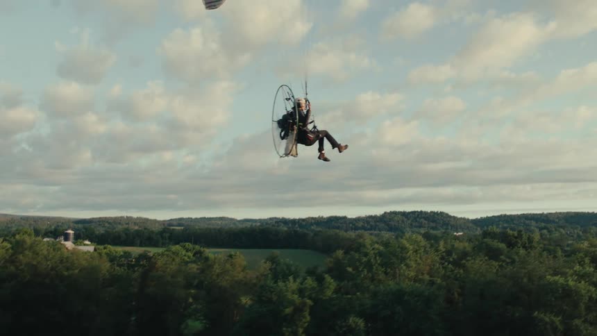 A powered paraglider soars gracefully in slow motion over a lush, green rural landscape during a beautiful sunrise
