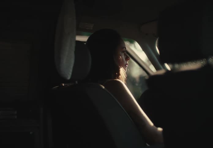A pensive young woman sits in the passenger seat of a car, her face illuminated by the warm, late afternoon sunlight as she gazes out the window