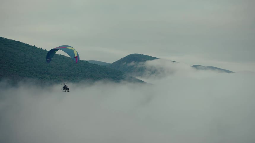 A powered paraglider slowly glides over a thick blanket of clouds covering a mountain valley
