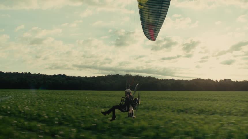 Cinematic side view of a paramotorist gliding smoothly over a vibrant green countryside field at golden hour
