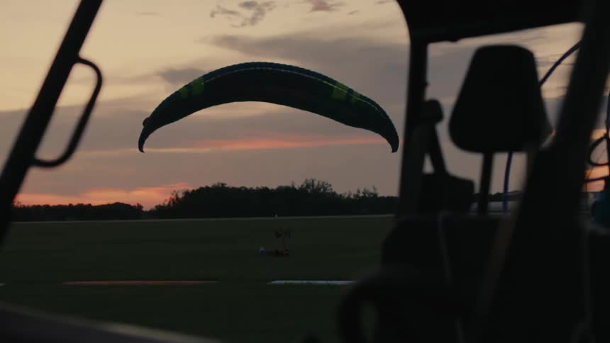 A powered paraglider's chute is inflated and ready for takeoff in a grassy field at sunset, framed by the interior of a vehicle