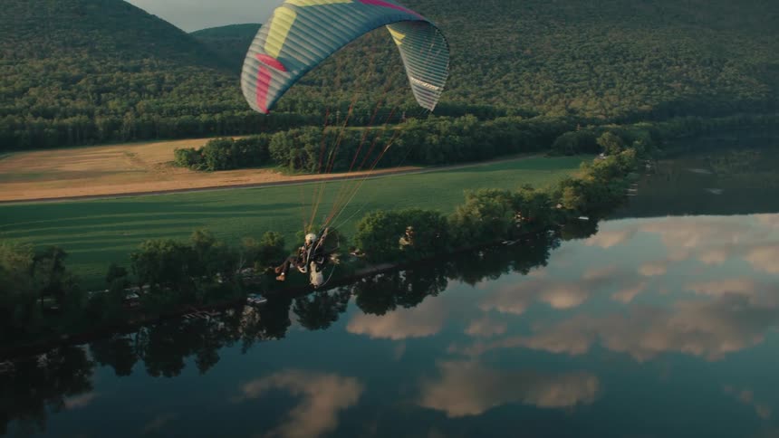A powered paraglider soars peacefully over a reflective river surrounded by lush green fields and forested hills