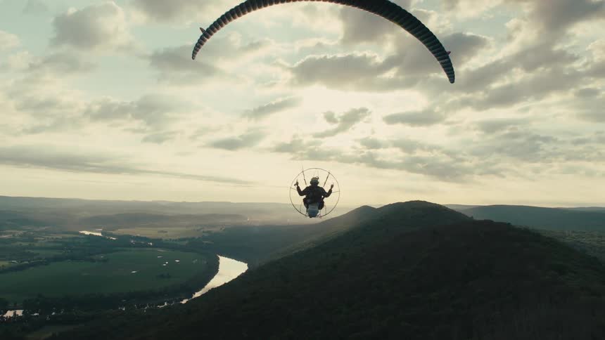 Cinematic slow motion shot of a paramotor pilot silhouetted against a golden hour sky, soaring over a mountain peak and a winding river valley