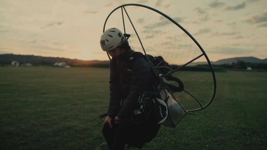 A powered paraglider pilot starts his motor and begins his takeoff run across a grassy field in slow motion at sunset