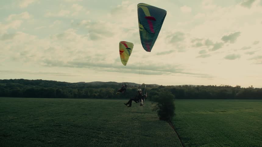 Two paramotorists fly in slow motion over lush green fields during a beautiful golden hour sunset