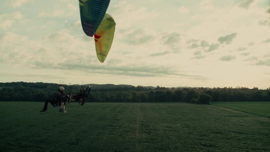 Two adventurous paramotor pilots fly in slow motion over a vast green field during a beautiful sunset