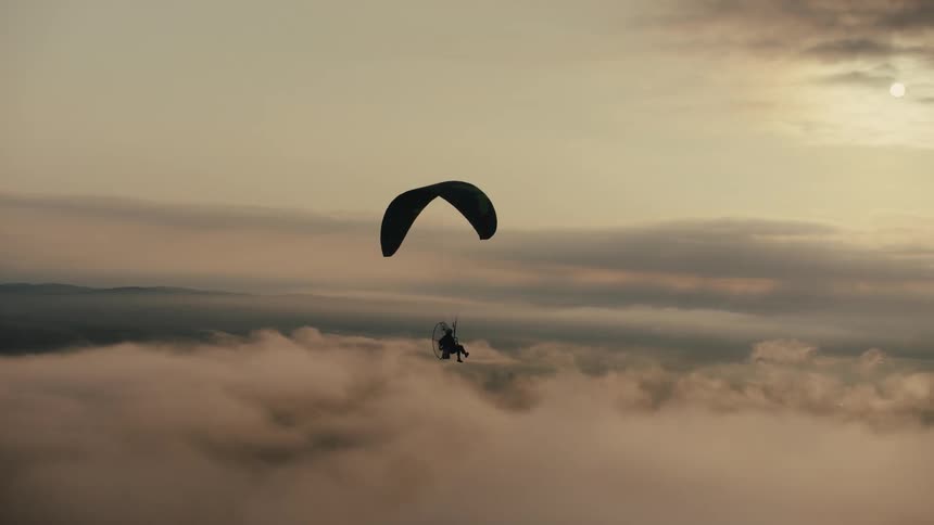 A powered paraglider soars peacefully in silhouette above a sea of clouds during a tranquil sunrise