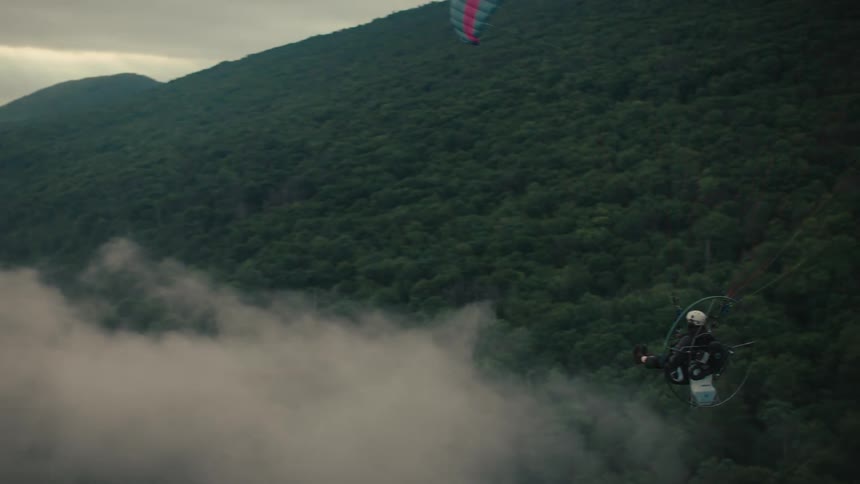 A person on a powered paraglider soars over a river winding through a foggy, tree-covered mountain landscape