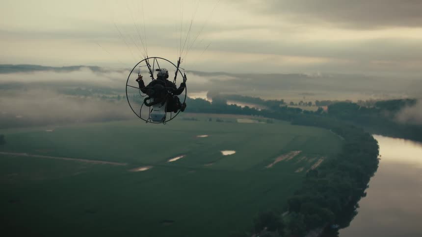 An epic aerial shot follows a powered paraglider flying high above a foggy river valley during a moody sunrise
