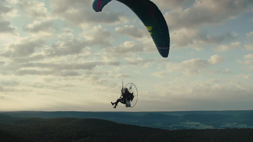 A slow-motion aerial shot follows a powered paraglider flying peacefully over rolling green hills under a cloudy sky at sunset