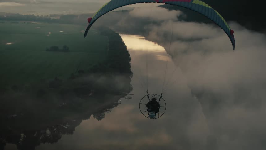 A person on a powered paraglider flies serenely through the clouds over a reflective river during a beautiful sunrise