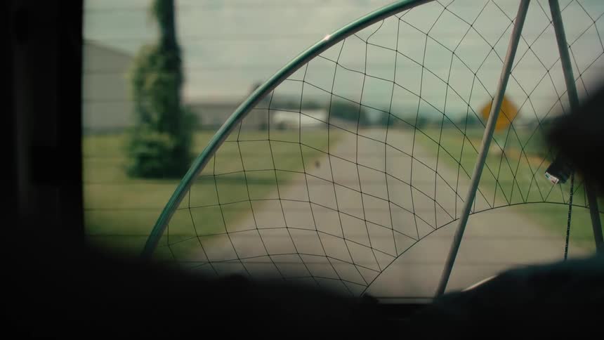 A view from the back of a moving car shows a paramotor frame resting against the window, with a country road and hangar in the background