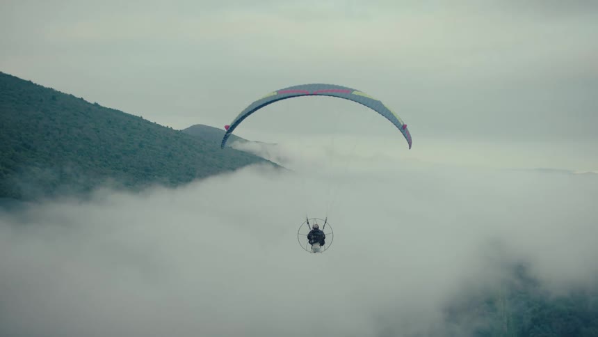 A powered paraglider glides through the sky, flying over a thick blanket of fog in the mountains