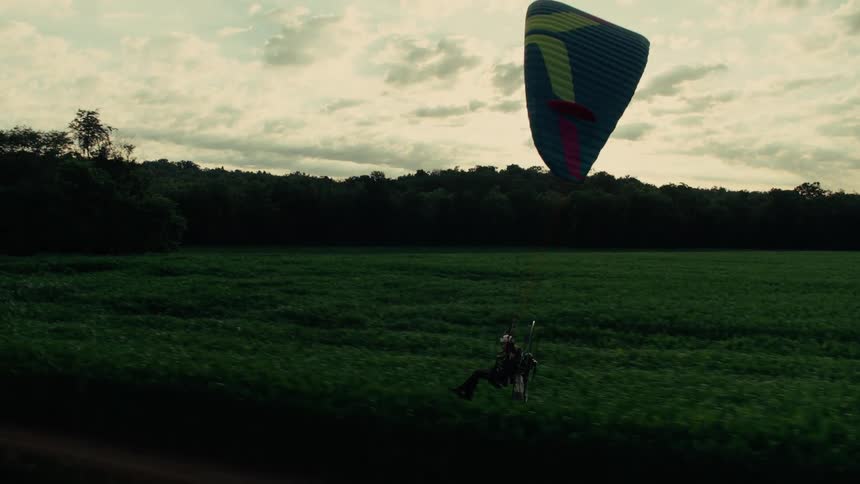 A person in a powered paraglider gracefully glides in slow motion over a lush green field before coming in for a landing