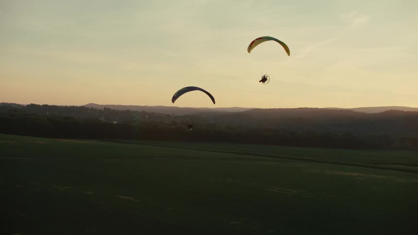Two powered paragliders soar gracefully over a vast green landscape during a stunning golden hour sunset