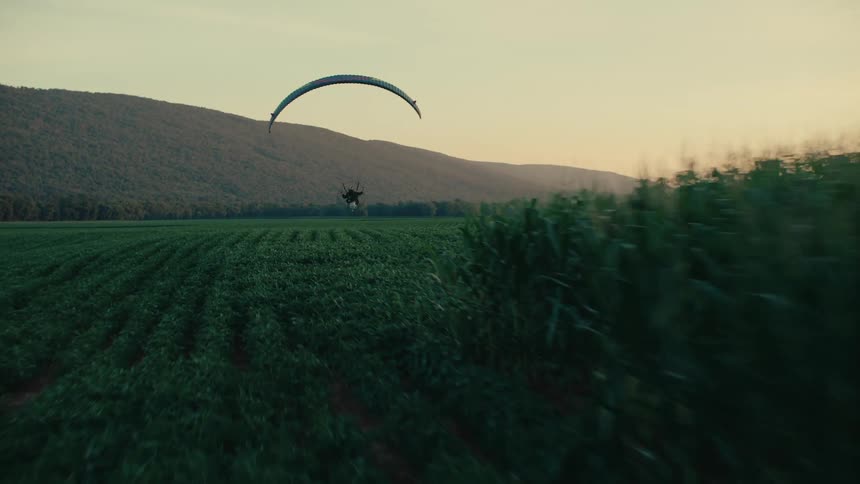 A powered paraglider soars low over a lush green farm field during a beautiful golden hour sunset