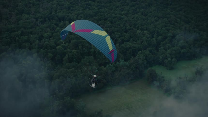 An aerial view captures a motorized paraglider soaring in slow motion over a vast, foggy forest