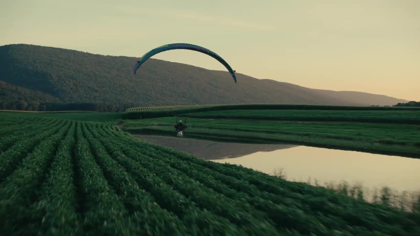 A powered paraglider glides low over vibrant green farm fields and a reflective pond during a beautiful sunset