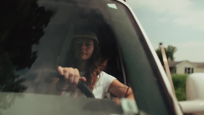 A young woman wearing a backward cap drives a van down a country road, seen from the front