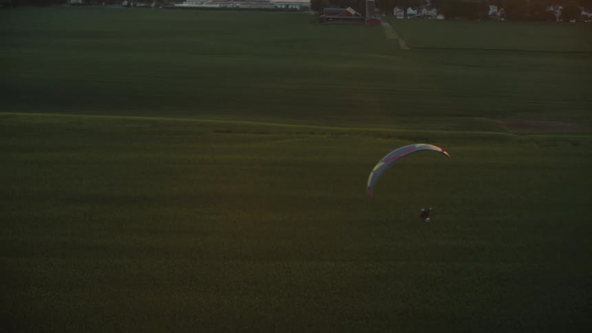 An aerial view of a person on a powered paraglider flying low over a vast green farm field at sunset