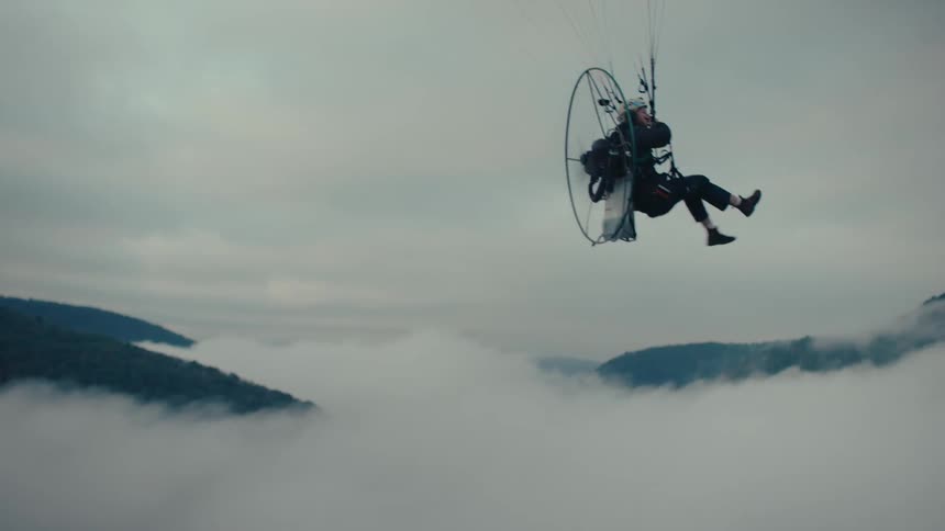 A person soars through the sky in a powered paraglider in slow motion, flying high above a thick layer of clouds in a mountain valley