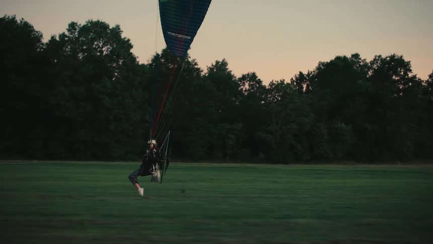 A powered paraglider pilot runs across a large grassy field to gain momentum for takeoff at dusk in slow motion