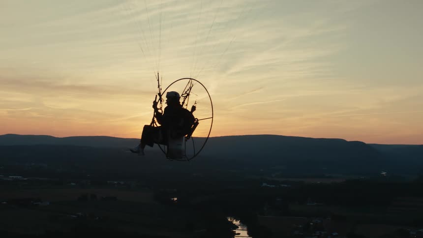 A silhouetted powered paraglider soars majestically over a rolling landscape as the sun sets on the horizon