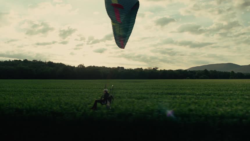 Side-view of a person on a powered paraglider flying low over a vibrant green field on a cloudy day