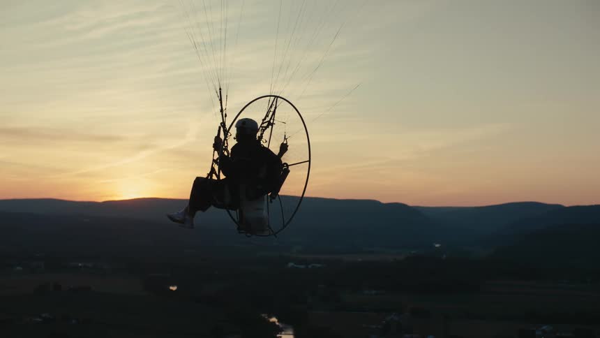 A silhouetted person enjoys a peaceful powered paraglider flight over a scenic mountain valley during a beautiful sunset