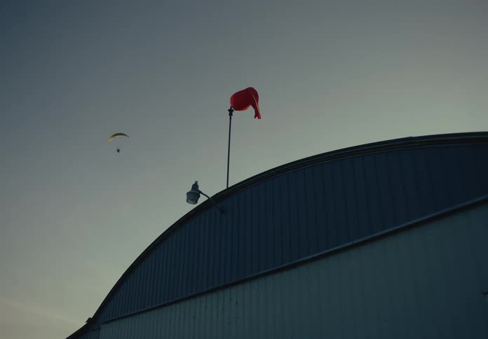 A static shot of a red windsock on top of an airplane hangar at dusk as a paraglider flies in the background