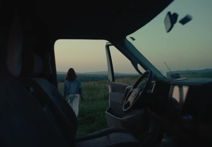 A solo female filmmaker exits her van with a vintage camera and walks into a scenic field to film at dusk