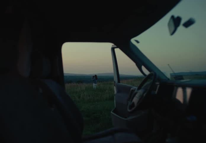 View from inside a van of a romantic couple hugging in a grassy field overlooking distant hills at dusk