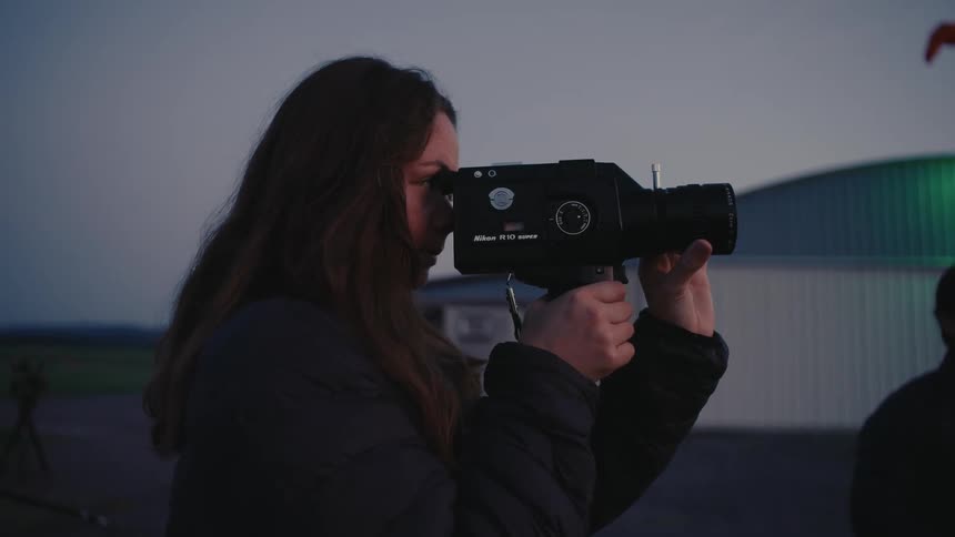 A female filmmaker carefully frames a shot with a vintage Nikon Super 8 camera at an airfield during twilight
