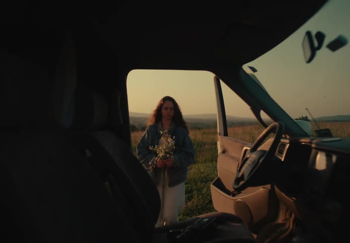 A young woman with a bouquet of wildflowers returns to her van in a serene field during a beautiful golden hour sunset