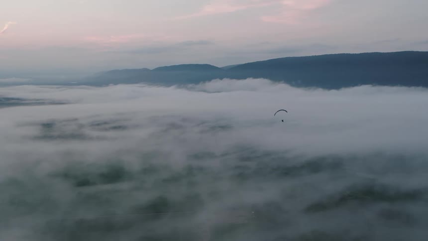 An epic aerial shot captures a paraglider soaring peacefully over a thick blanket of clouds in a mountain valley at sunrise