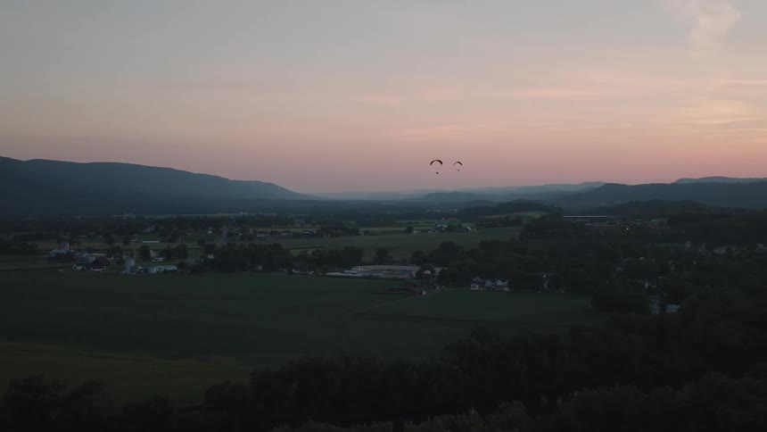 An aerial view shows two paragliders soaring gracefully over a rural mountain valley during a serene sunset