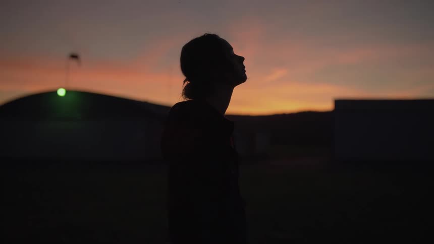 A silhouette of a contemplative woman slowly looks up at the colorful sunset sky