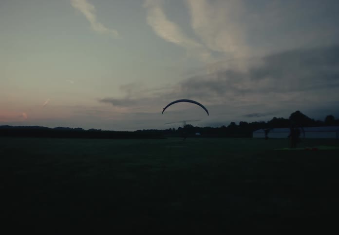A paramotor pilot takes off from a grassy field at dusk as another pilot prepares their gear