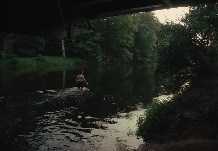 A young woman slowly walks into a calm river from under a bridge and begins to swim as twilight settles