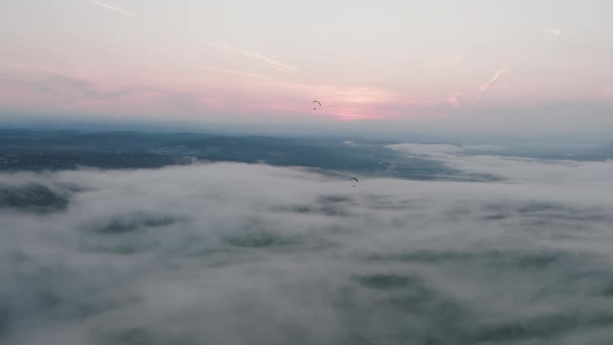 Aerial view of paragliders soaring peacefully above a sea of clouds covering a valley during a beautiful pink sunrise